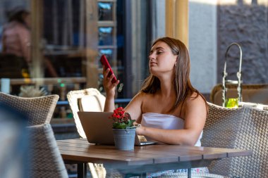 Genç kadın dizüstü bilgisayarda çalışıyor ve telefon kullanıyor sokak kafesinde oturuyor, uzaktan öğrenme ve çalışma konsepti