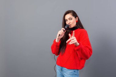 Genç, duygusal esmer, kırmızı kazak giymiş bir mikrofonla karaoke söylüyor ya da gri üzerine izole edilmiş bir konuşma yapıyor.