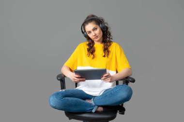 Sarı tişörtlü, uzun dalgalı saçlı bir genç. Bir ofis sandalyesinde oturuyor ve tablet kullanıyor, oyunlar oynuyor, müzik dinliyor. Gri renkte izole edilmiş.