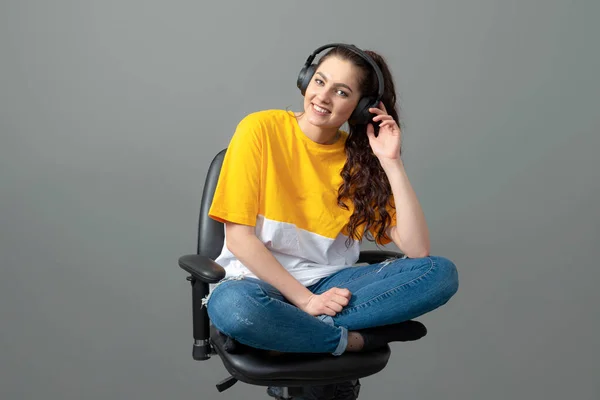 teenager with long wavy hair dressed in a yellow t-shirt sitting on an office chair and listening music, play games, listen to music, isolated on gray