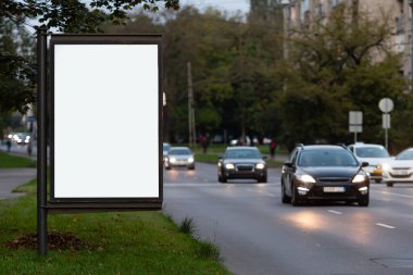 Şehir caddesinde dikey boş ilan panosu, arka planda arabalarla dolu gece sokağında.