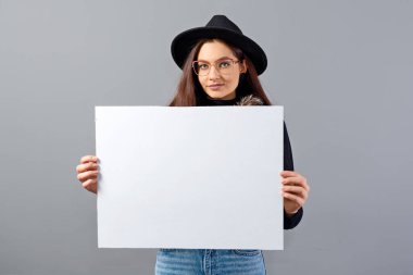 Gözlüklü, şapkalı, boş pankart gösteren zarif genç bir kadın. Gri renkte izole edilmiş, kopyalama alanı.