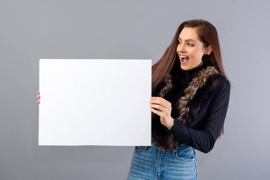 Şaşırmış genç bir kadın, gri üzerinde izole edilmiş, fotokopi alanı ile boş tabela gösteriyor.
