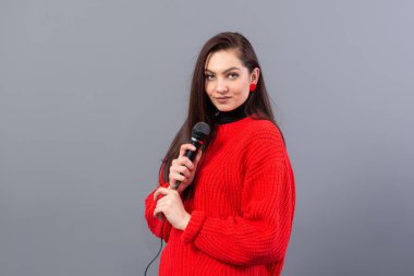 Genç, duygusal esmer, kırmızı kazak giymiş bir mikrofonla karaoke söylüyor ya da gri üzerine izole edilmiş bir konuşma yapıyor.