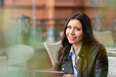 Sokak kafeteryasında oturmuş kameraya bakarken tabletle çalışan genç bir kadın.
