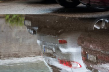 Kaldırımdaki su birikintisi, park halindeki bir arabanın yansıması, yakın plan, yağmur sonrası yol.