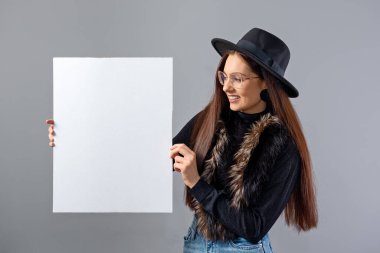 Gözlüklü, şapkalı, boş pankart gösteren zarif genç bir kadın. Gri renkte izole edilmiş, kopyalama alanı.