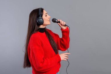 Genç, duygusal esmer, kulaklıklı ve kırmızı kazaklı bir mikrofonlu. Gri renkte karaoke söylüyor.