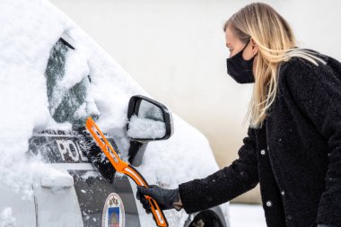 Yüz maskeli sarışın bir kadın bir polis arabasından kar temizliyor.