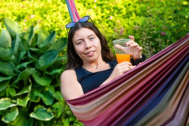 Harika kız hamakta uzanmış portakal suyu içiyor, bahçede dinleniyor.