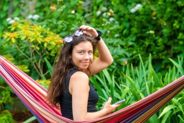 Güneş gözlüklü genç bir kız hamakta oturuyor, rahatlıyor ve akıllı telefon kullanıyor.