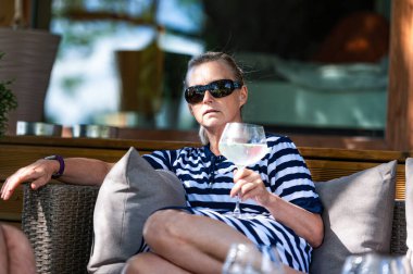 Orta yaşlı, güneş gözlüklü, elinde bir kokteyl ile bahçede dinlenen sarışın kadın.