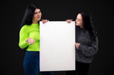 İki çekici, beyaz, boş posterli genç kadın, karanlık arka planda izole edilmiş, boşluğu kopyalıyorlar.