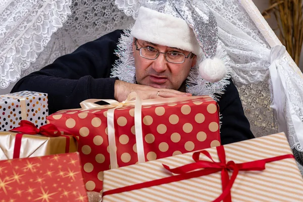 middle-aged man with glasses and a silver dwarf hat laying between christmas gift boxes in a decorated room, Christmas concept