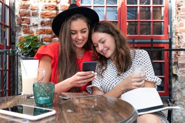İki neşeli genç bayan kahve içiyor, konuşuyor ve açık kafede akıllı telefon kullanıyor.