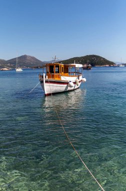 Tahta bir tekne turkuaz sularda dinleniyor, temiz bir gökyüzünün altındaki kayalık adalarla çevrili..