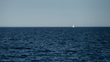 Küçük bir yelkenli, açık bir gökyüzünün altında derin mavi sularda süzülür..