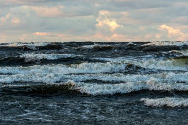 Gün batımında bulutlu bir gökyüzünün altında dalgalar sahile çarpıyor.