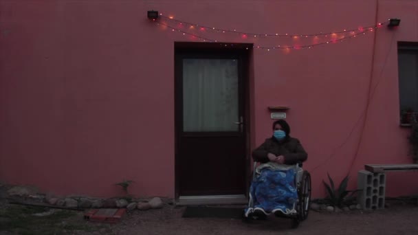 크리스마스등을 켜고 집 앞에서 건강에 좋은 모자를 쓰고 휠체어를 타고 있는 노인 — 비디오
