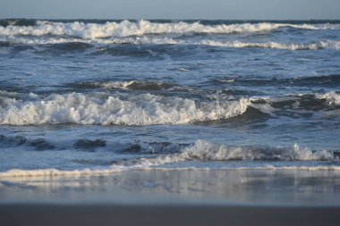 Gün batımında kumlu sahilde dalgalar yavaşça çarpıyor.