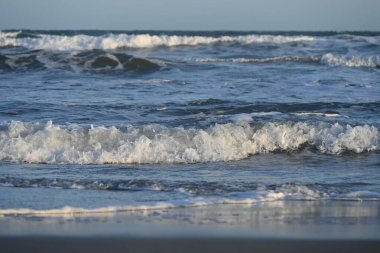 Dalgalar, açık mavi gökyüzünün altında, kumlu kumsalda yavaşça çarpıyor.