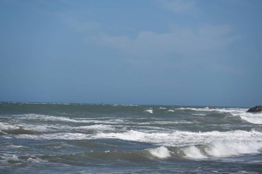 Dalgalar mavi gökyüzünün altında kayalık kıyılara çarpıyor.