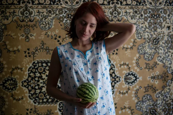Rothaarige Kaukasierin im Chintz-Nachthemd Nachtwäsche hält kleine Wassermelone mit dem sowjetischen Teppich im Hintergrund — Stockfoto