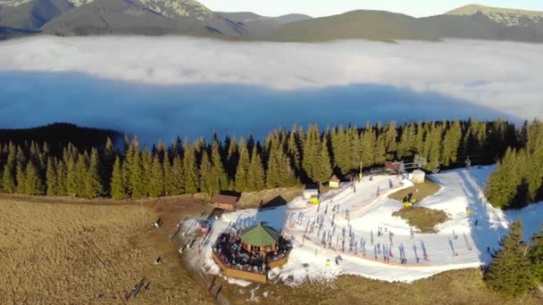 Flyg panorama över Bukovel Ski Resort berget Dovha, särskilt täckt med en konstgjord snö. Låga moln hänger runt berget och människor på himlen och Snowdoards njuter av solnedgången i ovanifrån — Stockvideo