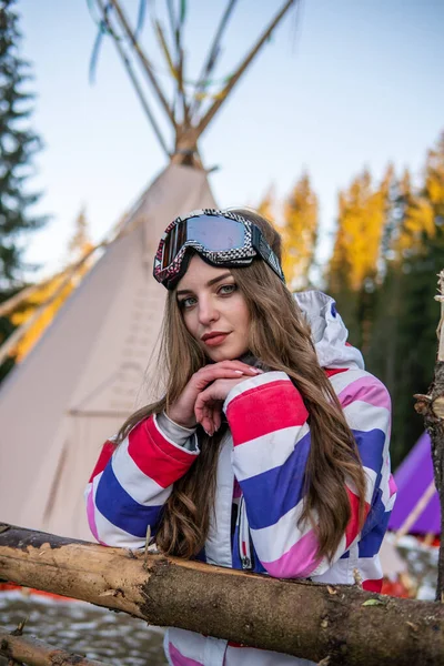 Porträt einer hübschen kaukasischen jungen Frau in der Nähe von Tipi oder Wigwam in Skianzug und Wintersportmaske auf dem Kopf. Porträt einer fröhlichen blonden Frau im Skigebiet — Stockfoto