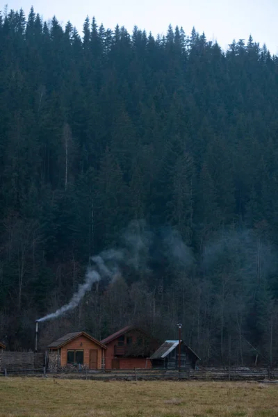 Fumo branco das chaminés de casas de madeira precisas em uma aldeia de montanha no outono em um fundo de floresta e montanhas — Fotografia de Stock