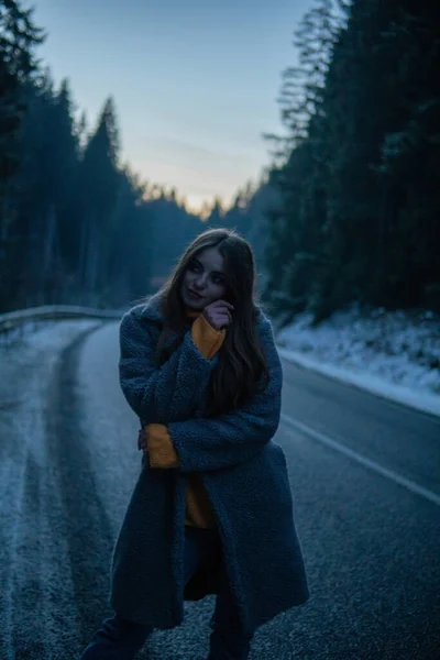 Belle jeune femme sur la route de la forêt sombre à l'hiver. Crépuscules, lumières de voiture sur le fond — Photo