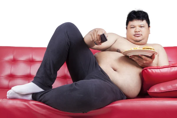 Ожирний чоловік дивиться телевізор, тримаючи бургер — стокове фото