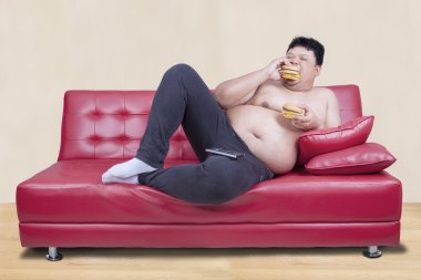 Obez adam kanepede eğilerek hamburger yiyor