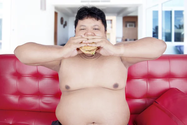 Homem com excesso de peso come hambúrguer no sofá — Fotografia de Stock