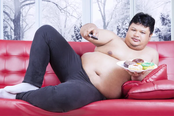 Homem com excesso de peso assistindo tv e come donuts — Fotografia de Stock