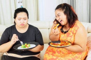 Kilo verme konsepti. Mutsuz şişman kadın salata yerken ablasının alaycı ifadesiyle pizza yiyor.