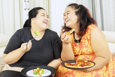 Oturma odasında kanepede otururken birlikte pizza yiyip salata yiyen iki şişman kadın. Evde vuruldu.