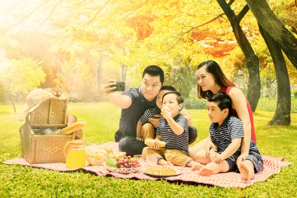 快乐的一家人一边在公园里野餐一边用手机给自己拍了一张秋树背景的自拍照片 — 图库照片