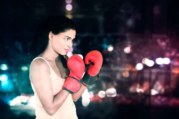 Side view of young woman ready to fight while wearing boxing gloves and standing with blurred lights background