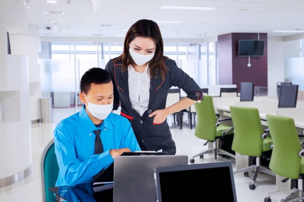 男性上司は医療マスクを着用しながら オフィスで彼の従業員と話し ラップトップコンピュータを使用しています — ストック写真