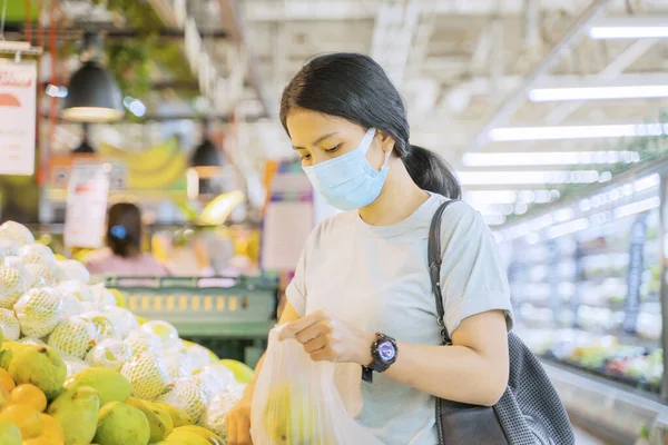 新しい通常の生活の中でスーパーマーケットで新鮮な果物を選択しながら顔マスクを身に着けている若い女性 — ストック写真