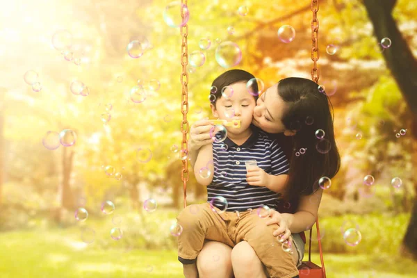 幸せな母親キス彼の息子ながら吹いて石鹸泡と一緒に座ってスイングで秋の木の背景 — ストック写真