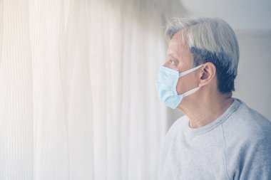 Evde karantina sırasında hayal kuran ve pencerenin yanında duran yaşlı bir adamın yüz maskesi takması.