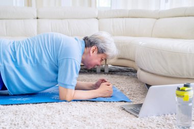 Üst düzey adam dizüstü bilgisayarda egzersiz videosu izlerken ve evde karantina yaparken tahta egzersizi yapıyor.