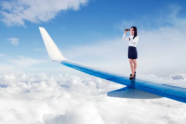 年轻的女商人站在飞机的机翼上 一边在云层上飞翔 一边用双筒望远镜看 — 图库照片
