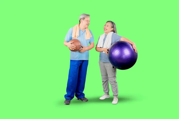スポーツウェアを身にまとい ピラティスボールとバスケットボールを持ち 緑のスクリーンを背景にスタジオに立つお年寄りカップルの全長 — ストック写真