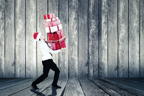 男性マネージャーは木製の壁の背景とサンタの帽子を身に着けている間クリスマスプレゼントの山を運ぶ — ストック写真
