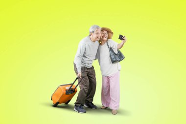 Happy old couple taking a selfie photo together by using a smartphone while carrying a luggage in the studio with green screen background