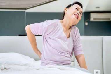Yatakta otururken uyandıktan sonra sırtı ağrıyan yaşlı bir kadın resmi. Evde vuruldu.