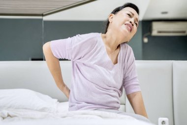 Picture of old woman having backache after waking up while sitting on the bed. Shot at home clipart